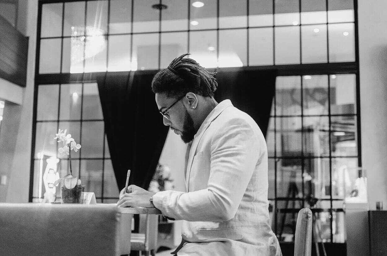 Corey writing at a table by tall windows, in black and white